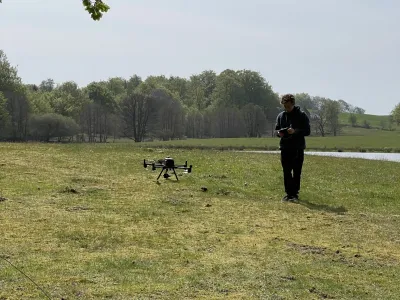 Person operating a drone in an open grassy field with trees and water in the background
